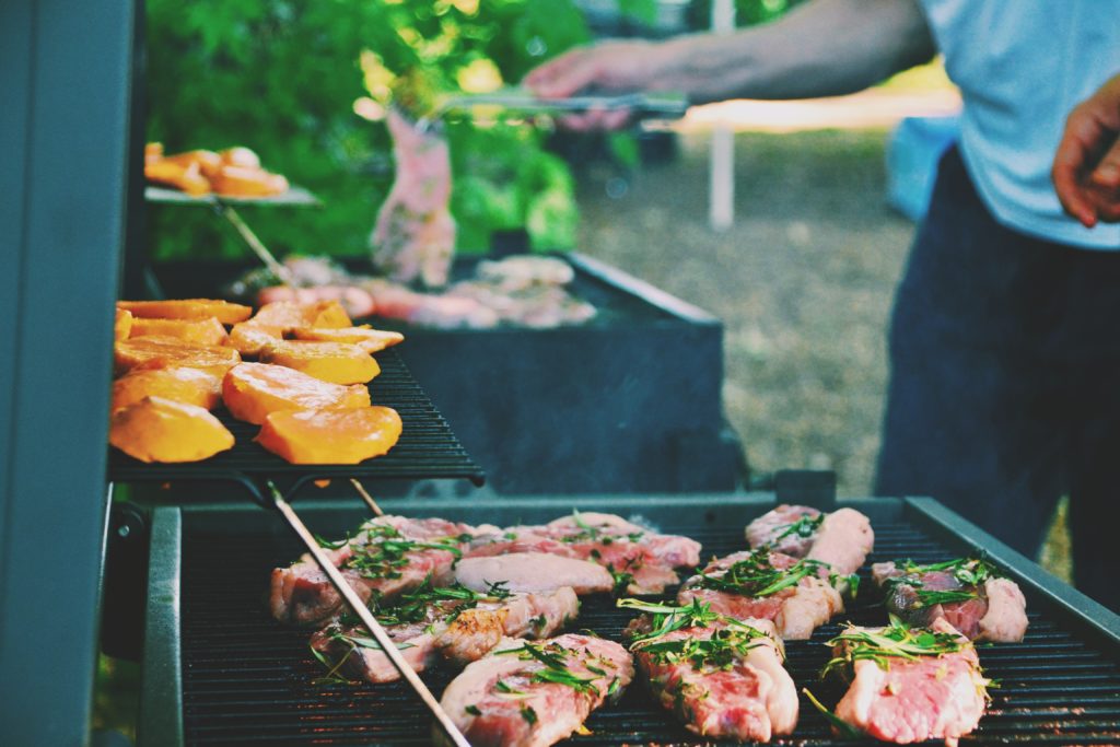 Man standing over his BBQ with a large selection of steak & chicken grilled with herbs on top.