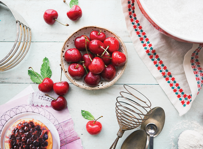 Cherries laying out on a kitchen counter among other cooking baking itesm, such as spoons, plates, towels