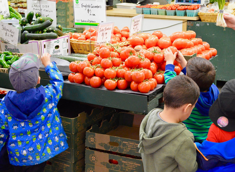Kids reaching out to touch the display of fresh produce inside the Hamilton Farmers' Market while completing the Scavenger Hunt